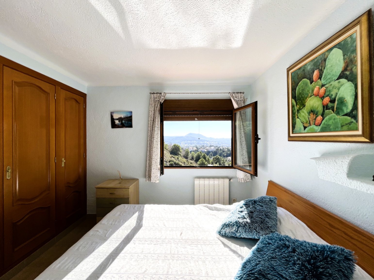 Bright bedroom featuring built-in wooden wardrobes, central heating radiator, and an open window offering expansive views of the Montgó mountain landscape.