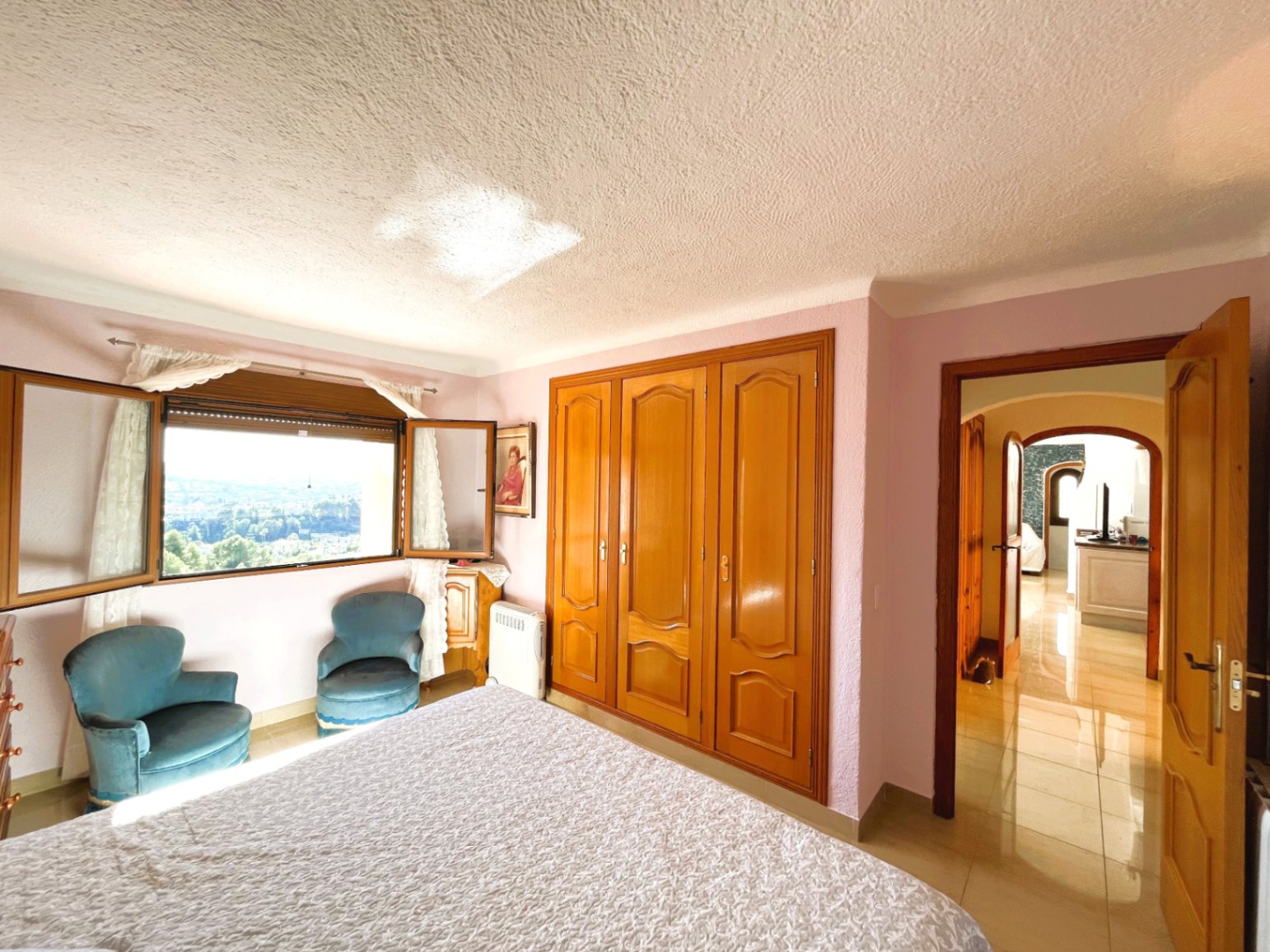 Bright bedroom featuring built-in wooden wardrobes, velvet armchairs, and a window offering expansive panoramic landscape views.