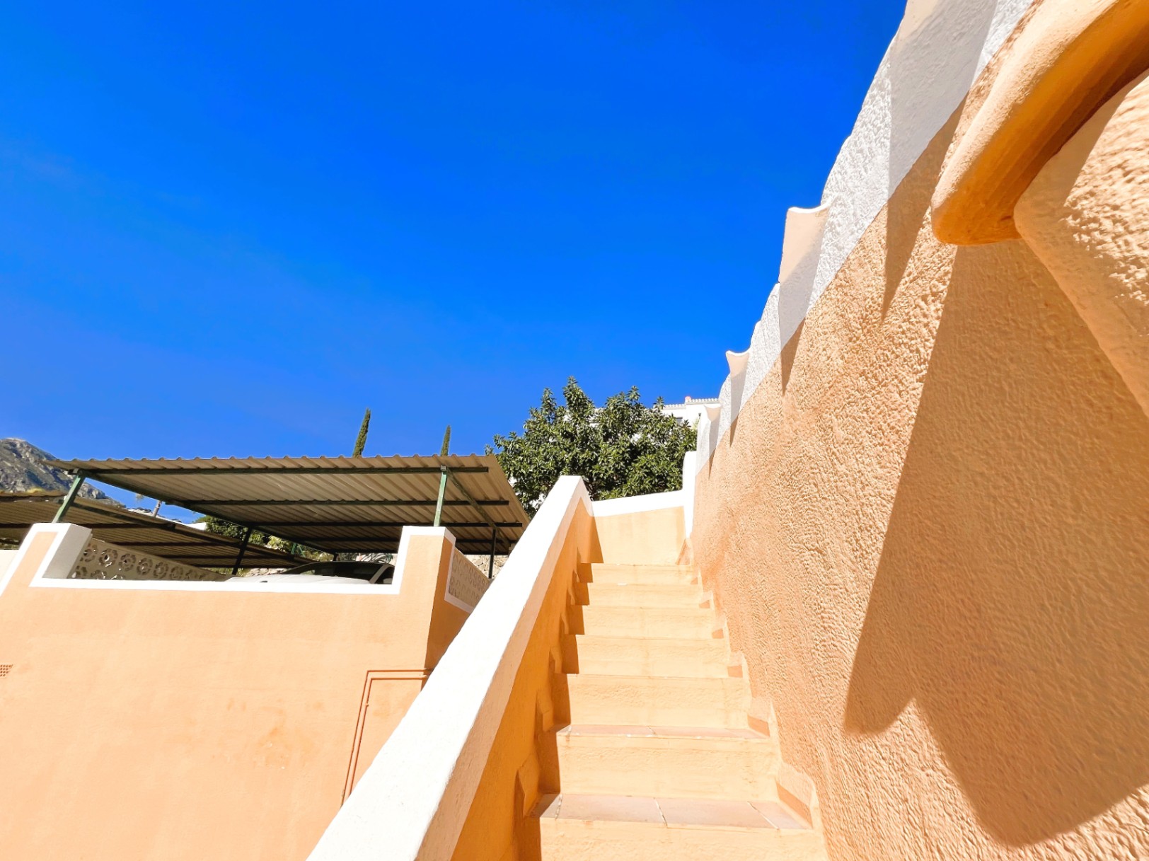 Exterior tiled staircase with terracotta-toned walls and white trim, located adjacent to the covered parking area with a metal pergola.