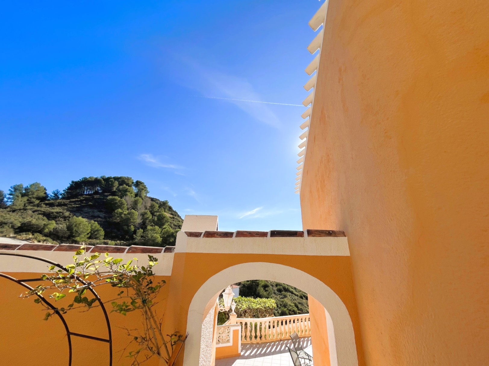 Exterior view featuring a white arched entryway against an ochre facade, with terracotta roof tiles and views of the pine-covered hills.