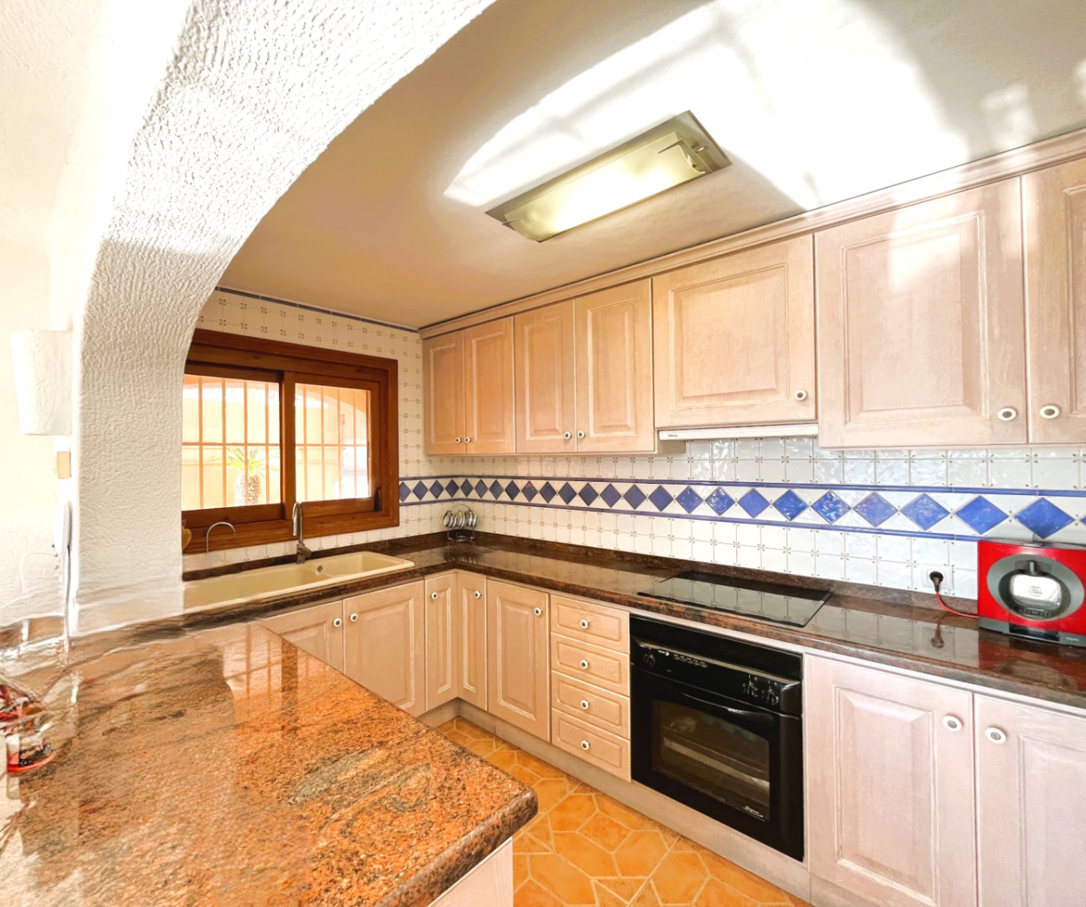 Kitchen featuring light wood cabinetry, granite countertops, and a tiled backsplash with blue accents. Includes an integrated oven, ceramic hob, and a rustic wooden window.