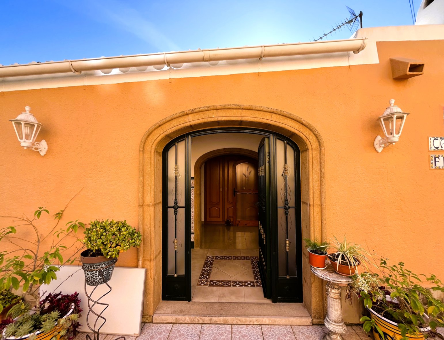 Main entrance featuring a natural stone arch, wrought iron security door with frosted glass, and ochre facade accented by white lanterns and potted greenery.