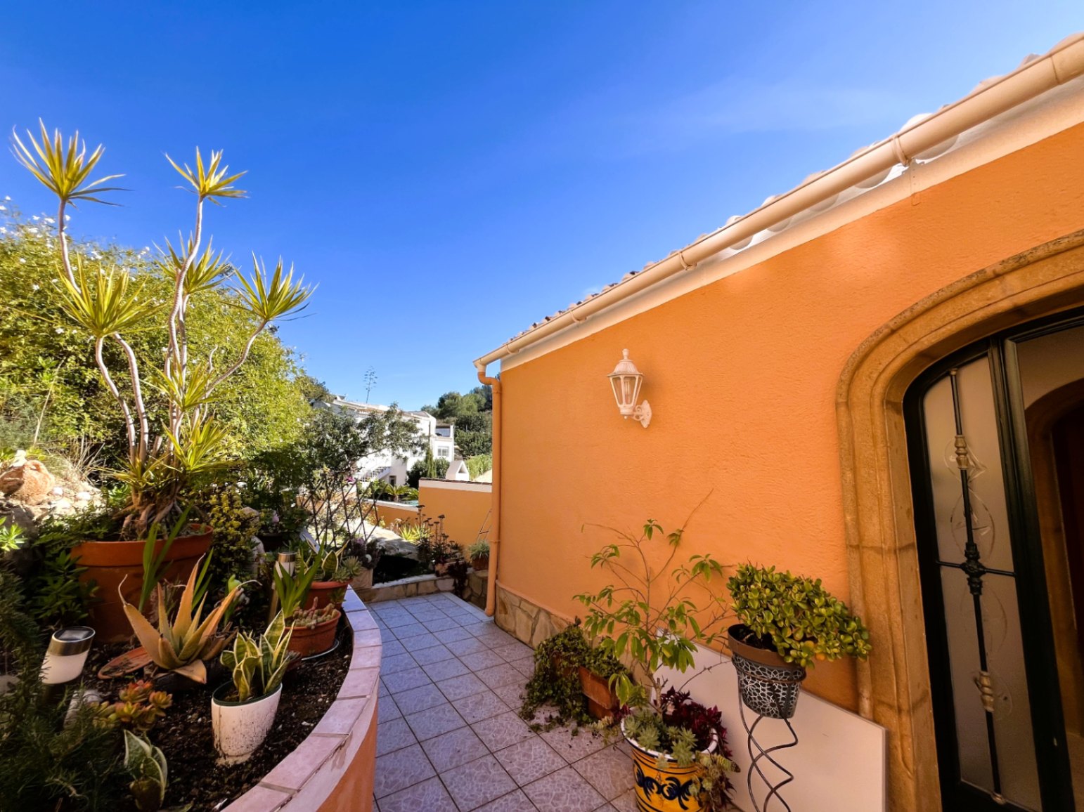 Outdoor tiled walkway featuring a curved planter with succulents, an ochre facade with a traditional stone archway, and a decorative wall lantern.