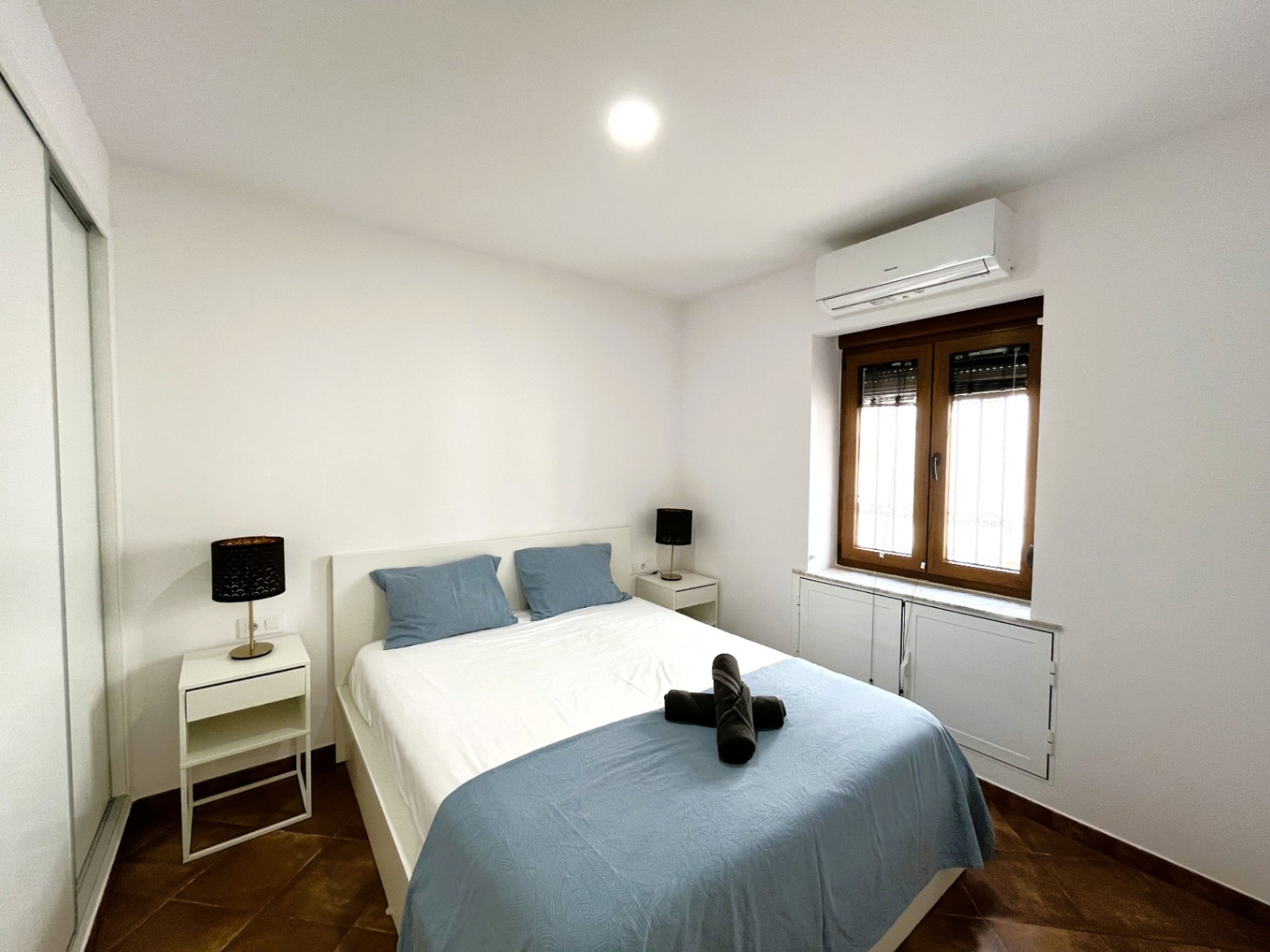 Bedroom featuring a double bed, white nightstands, and split air conditioning. Includes rustic tiled flooring, a built-in wardrobe, and a wood-framed window.