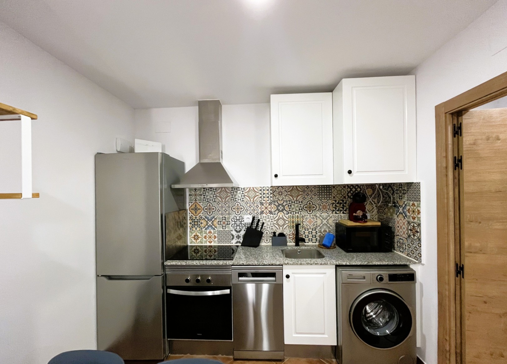 Compact renovated kitchen featuring a granite countertop, hydraulic-patterned tile backsplash, and integrated stainless steel appliances including a dishwasher and washing machine.