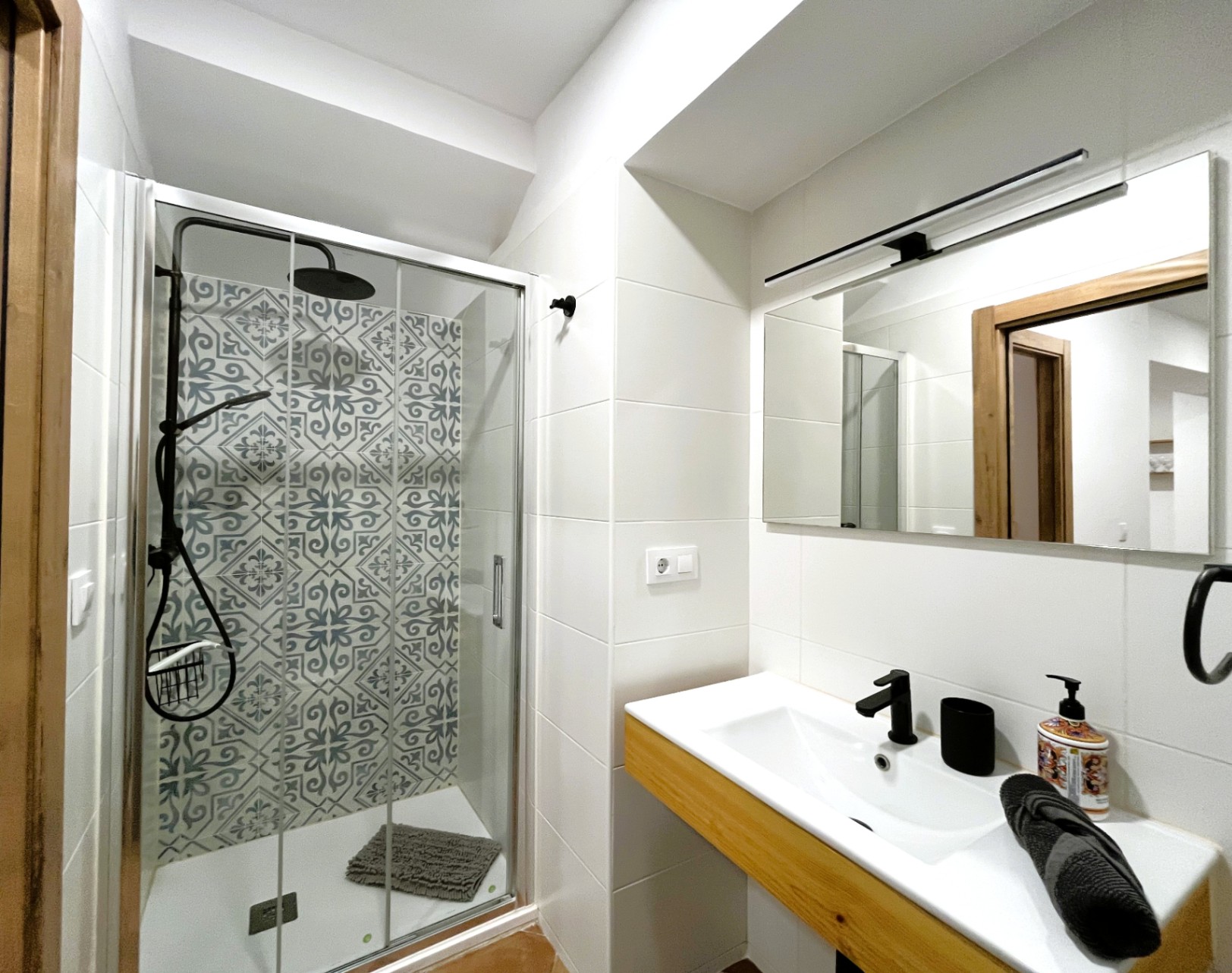 Modern bathroom featuring a walk-in shower with patterned tiles and black fixtures. A white vanity with wooden accents and a linear light above the mirror complete the space.
