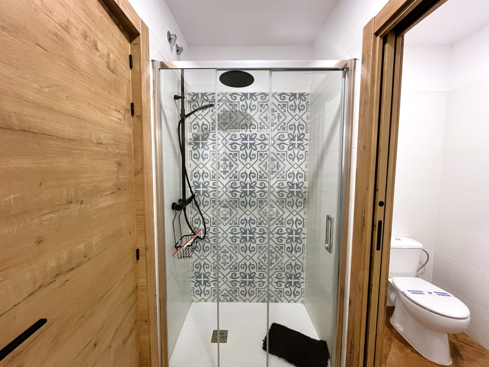 Renovated bathroom featuring a walk-in shower with glass doors, patterned hydraulic-style tiles, and black fixtures, framed by natural wood-finish doors.