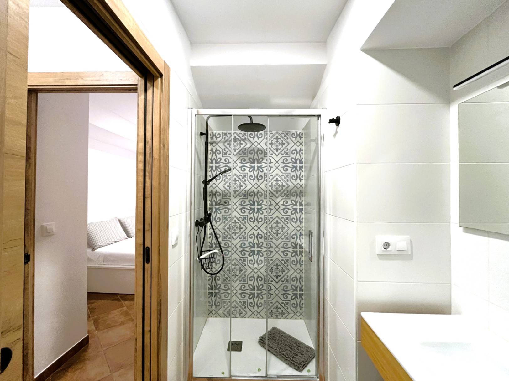 Renovated bathroom featuring a walk-in shower with patterned hydraulic tiles and black fixtures. View through the wooden doorway into the bright bedroom.