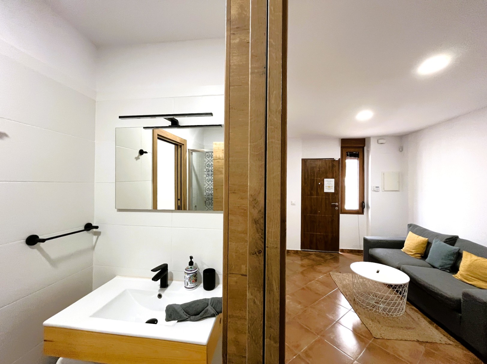 View of the living area with terracotta floors and grey sofa, adjacent to a modern bathroom featuring a white sink, black fixtures, and clean tiling.