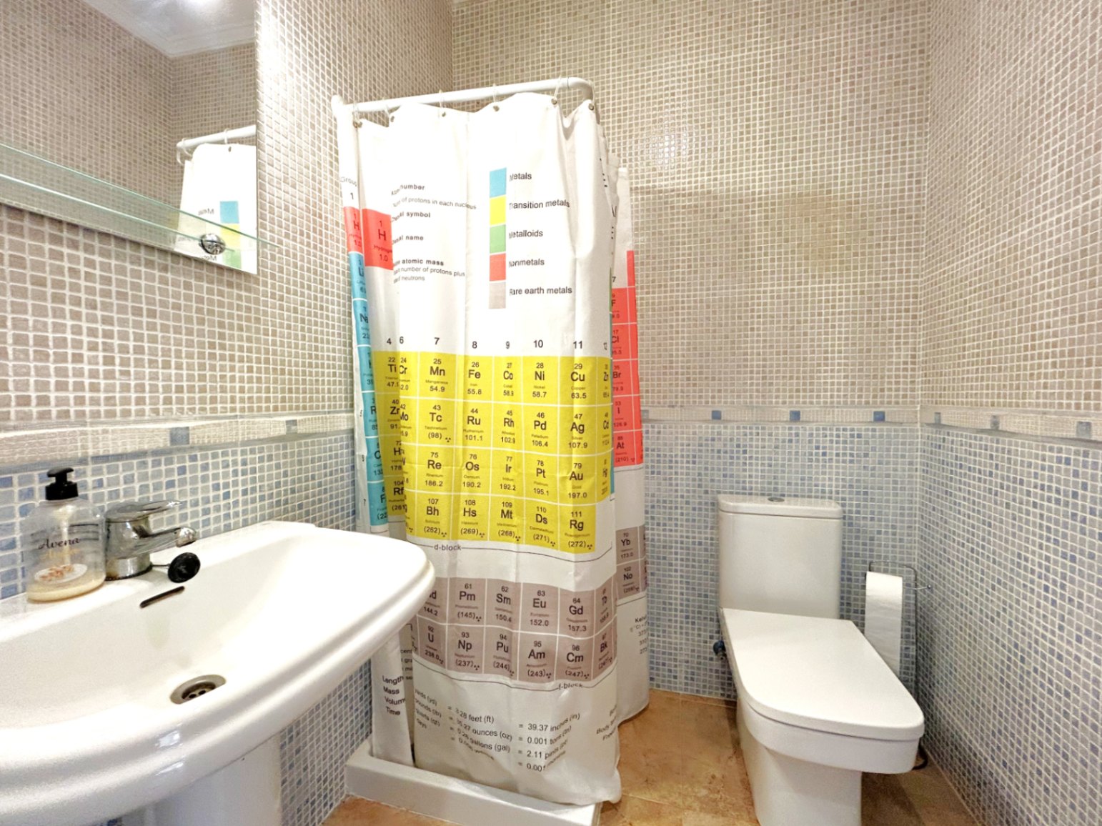 Bathroom featuring dual-tone mosaic wall tiling, a white pedestal sink, and a periodic table shower curtain. Includes a modern toilet and a mirror with a glass shelf.