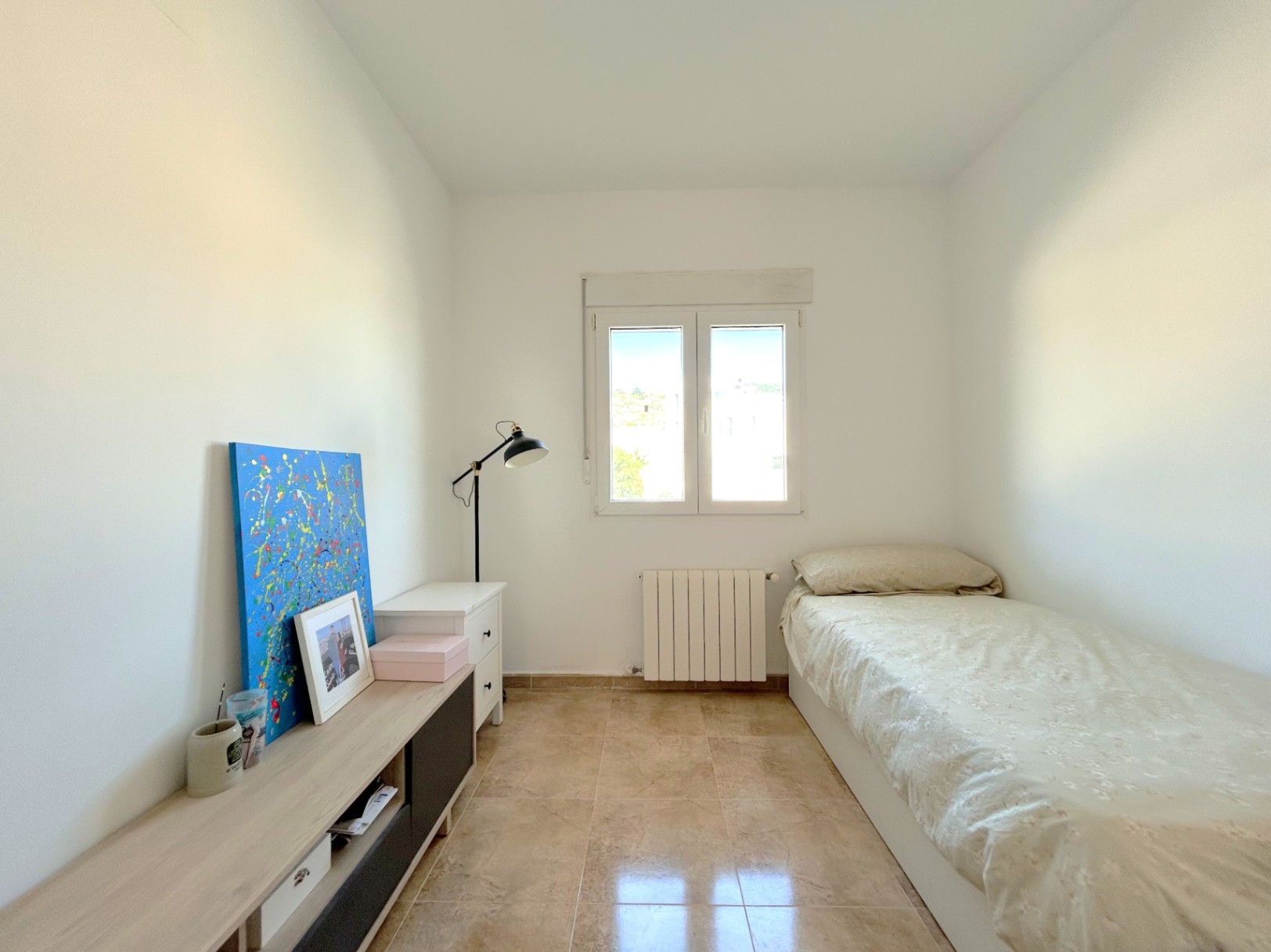 Bright bedroom featuring tiled flooring, a central heating radiator, and a double-pane window providing ample natural light.