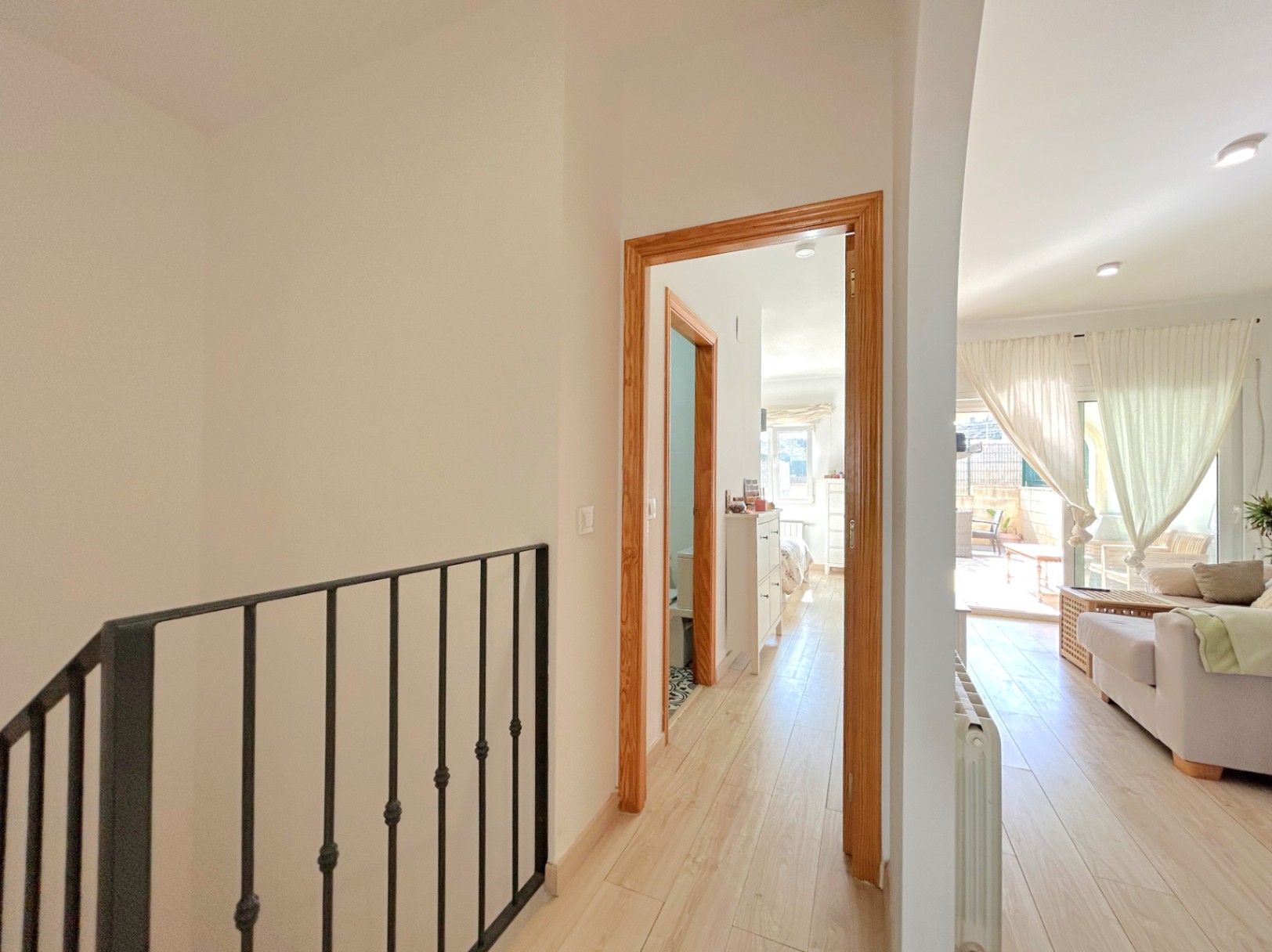 Bright hallway with light wood flooring and wrought iron railings, connecting the living area to the bedrooms and bathroom, featuring a central heating radiator.