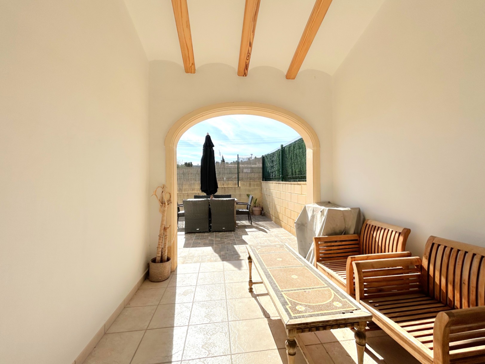 Covered terrace featuring exposed wooden ceiling beams, a classic archway, and tiled flooring, leading to a sun-drenched outdoor dining area.