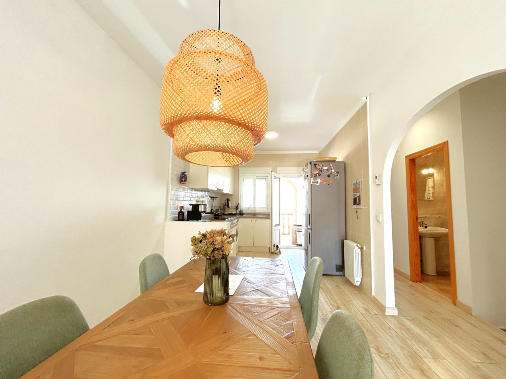 Dining area featuring a patterned wooden table and wicker pendant light, seamlessly connected to an open kitchen with white cabinetry and tiled walls.