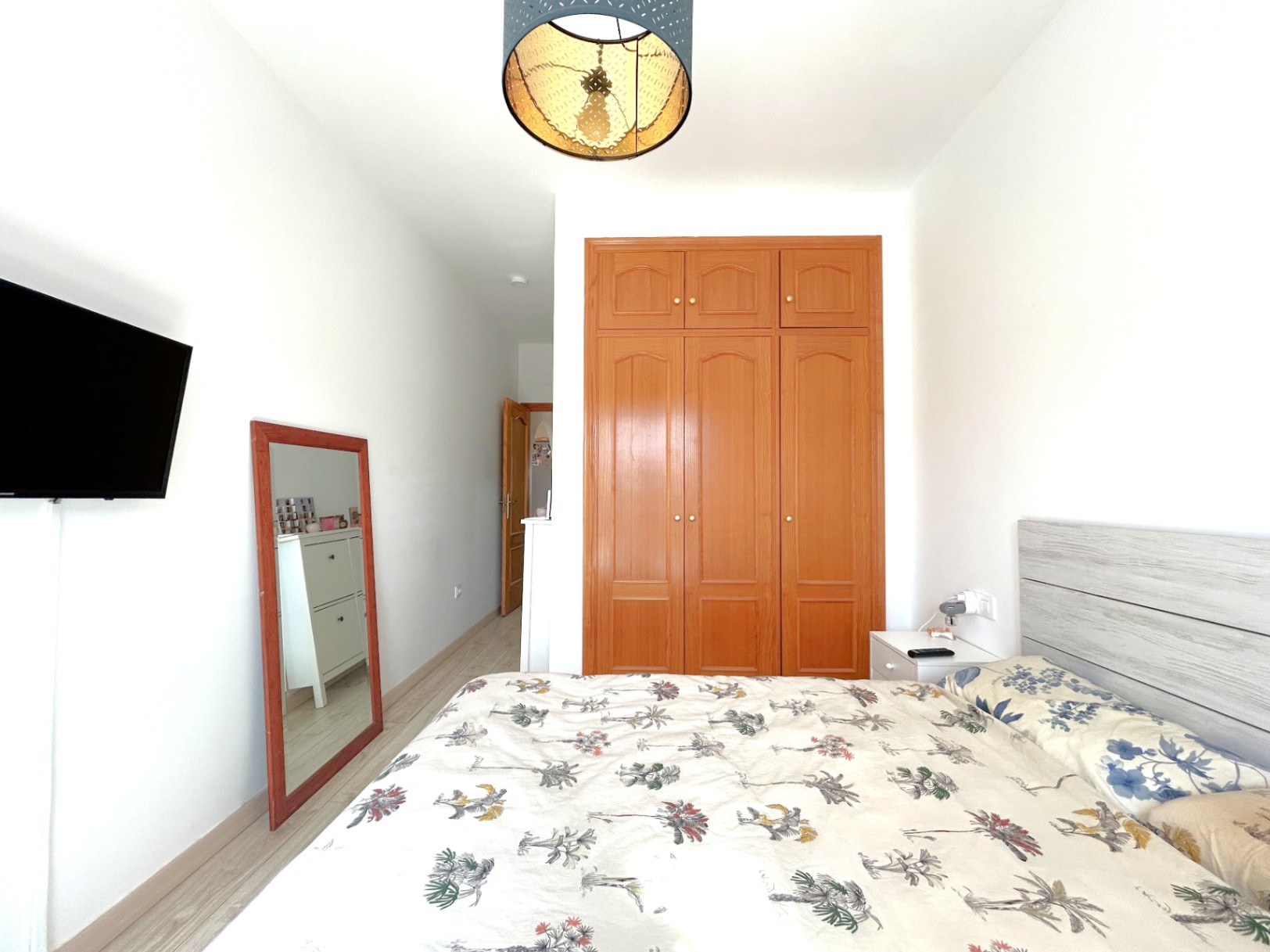 Double bedroom featuring a built-in wooden wardrobe, light laminate flooring, and a wall-mounted TV. It includes a full-length mirror and a decorative ceiling lamp.