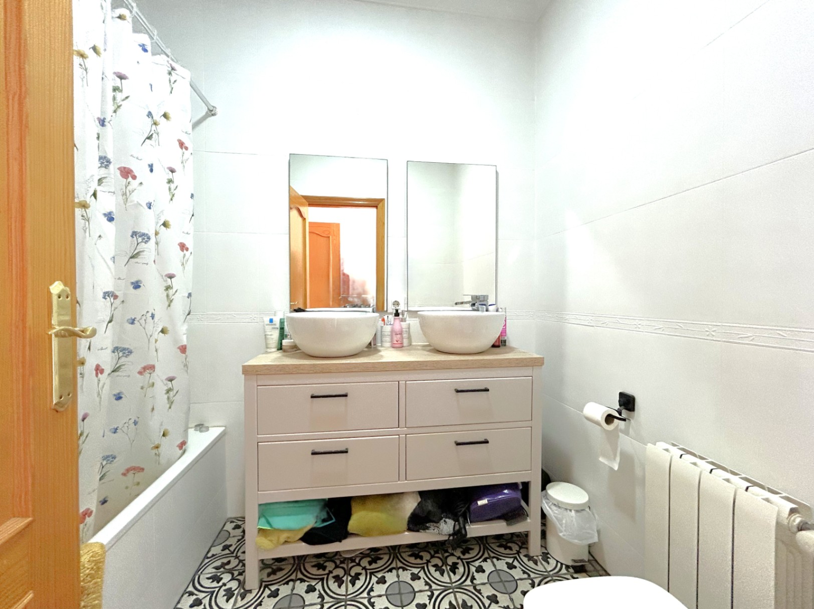 Full bathroom featuring a double vanity with vessel sinks, decorative hydraulic floor tiles, a bathtub with floral curtain, and a central heating radiator.