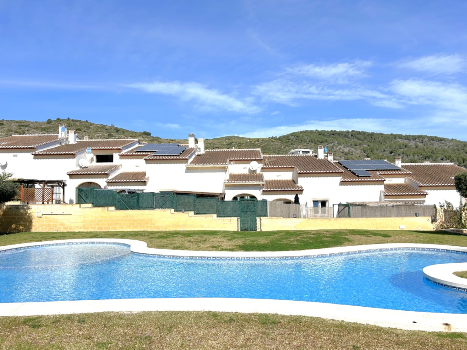 Large communal swimming pool with curved edges and lawn area, situated in front of Mediterranean-style townhouses with solar panels and mountain views.