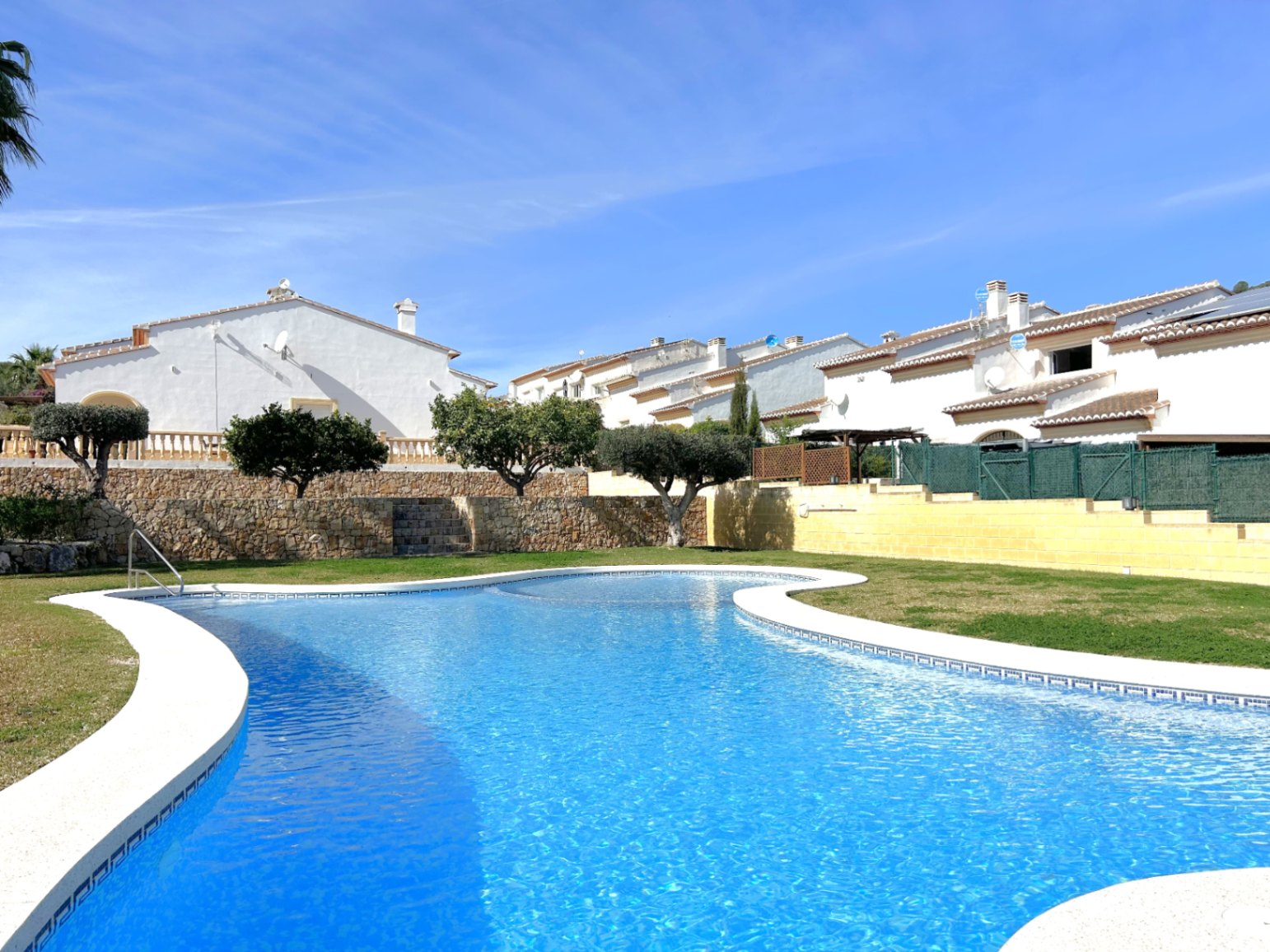 Large communal swimming pool with curved edges, bordered by a manicured lawn and natural stone walls against a clear sky.