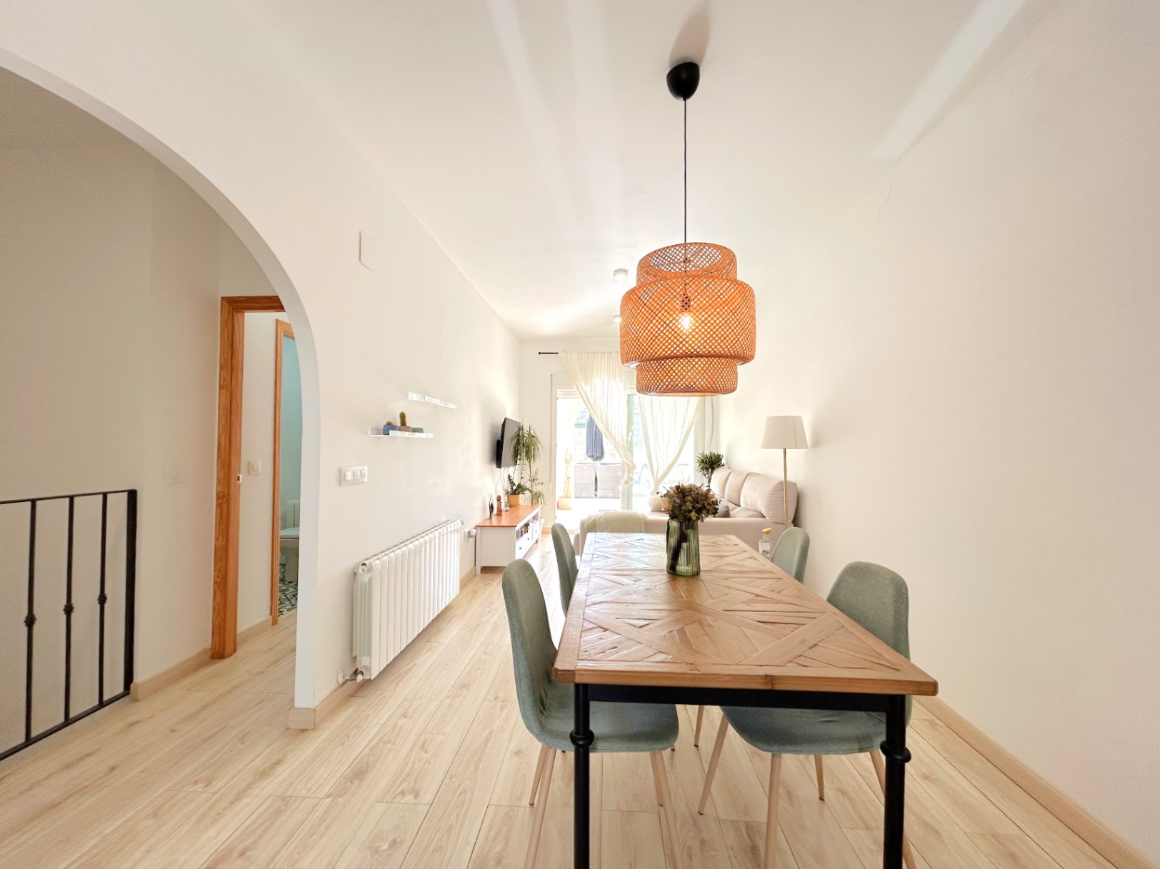Open-plan living area featuring a geometric wood dining table, wicker pendant light, and light flooring. The space includes central heating and leads to the sunlit terrace.