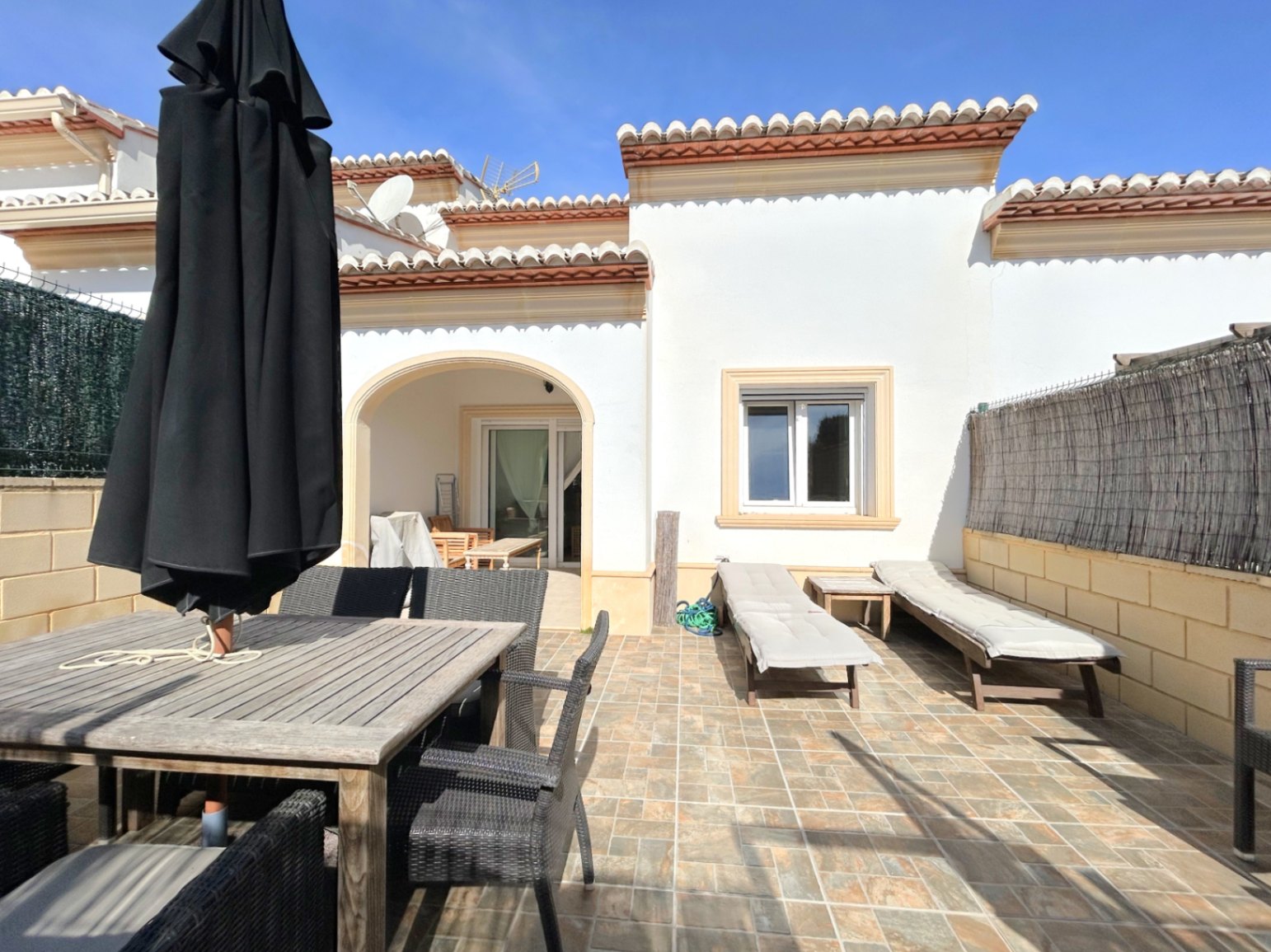 Private tiled terrace featuring a wooden dining set, sun loungers, and a shaded arched porch. The Mediterranean-style facade is finished with traditional roof tiles.