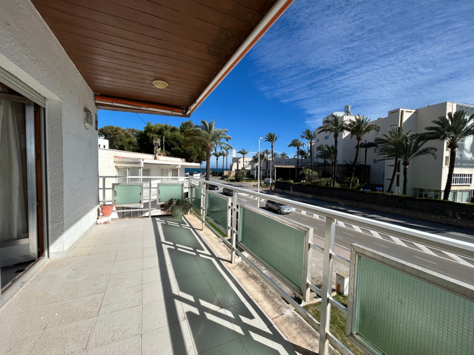 Outdoor terrace featuring ceramic tiling, a wood-paneled ceiling, and metal railings with glass inserts. It overlooks a palm-lined avenue near the coast.