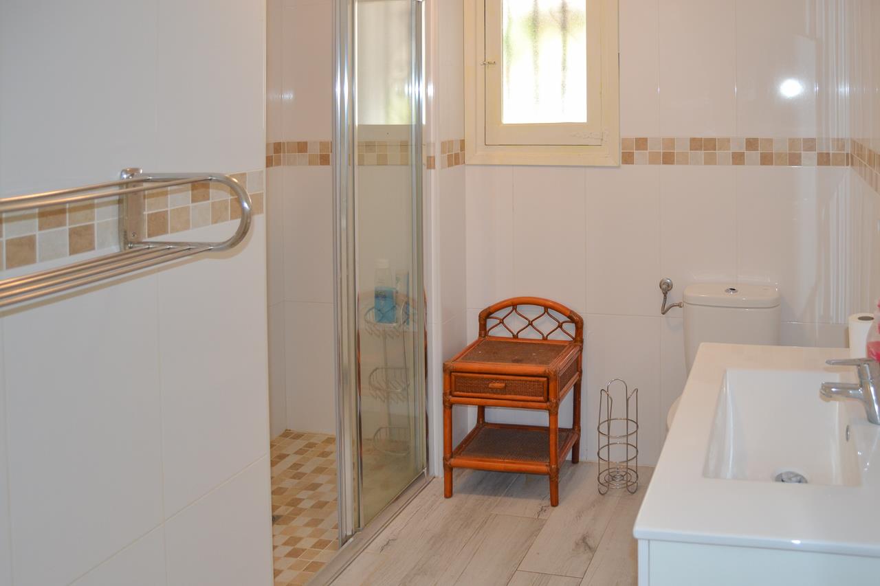 Bathroom with white tiled walls and mosaic borders. Features a glass shower enclosure, integrated sink vanity, and wicker side furniture under a frosted window.