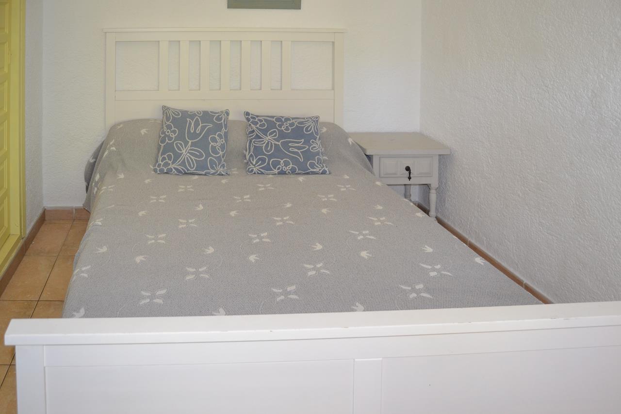 Bedroom featuring a white wooden bed frame with matching headboard and nightstand. The space includes textured white walls, terracotta floor tiles, and blue floral accent pillows.