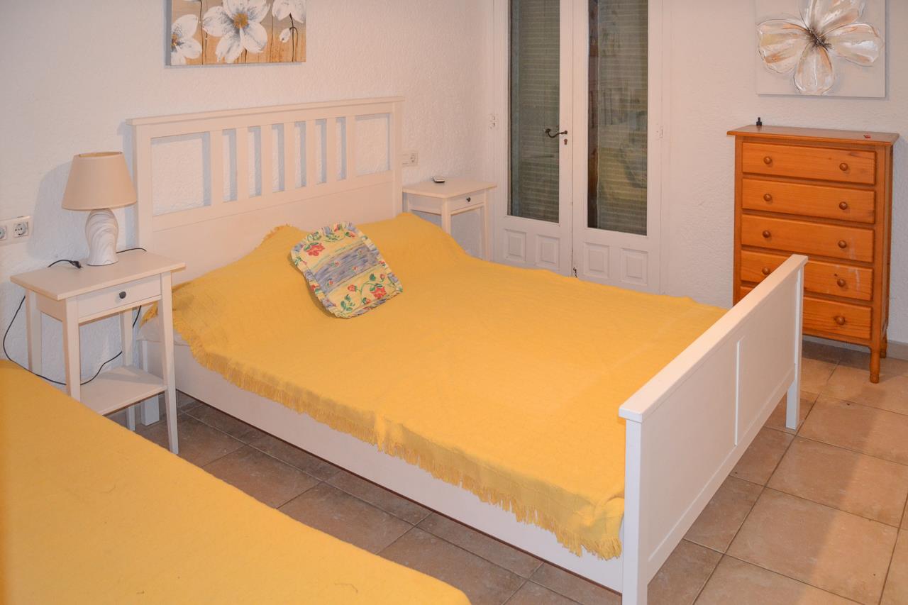 Bedroom featuring a white wooden bed frame with yellow linens, matching nightstands, and a pine chest of drawers. The space includes tiled flooring and glazed doors.