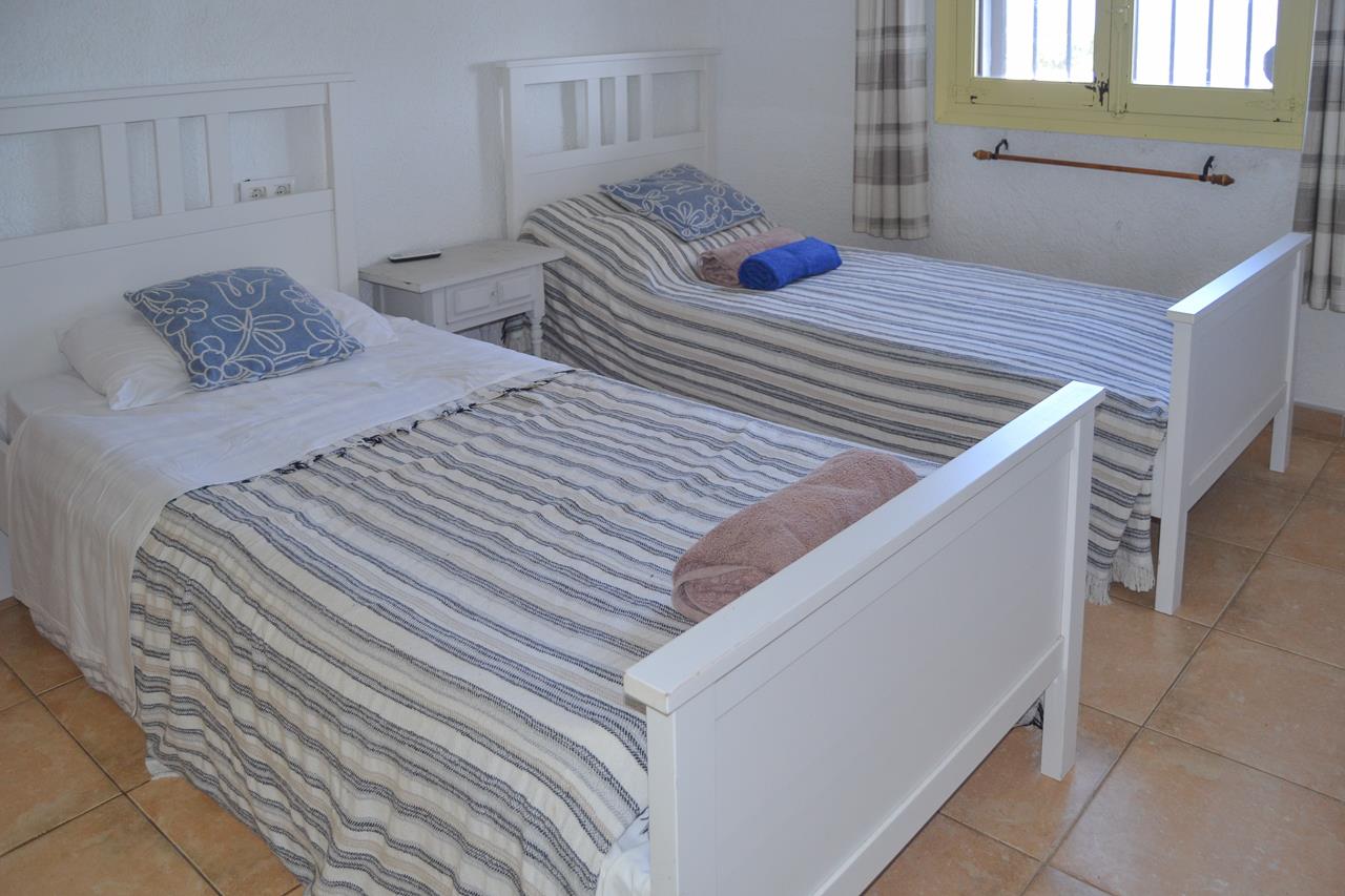 Bedroom featuring twin white wooden beds, rustic ceramic tile flooring, and a window with yellow frames and plaid curtains.