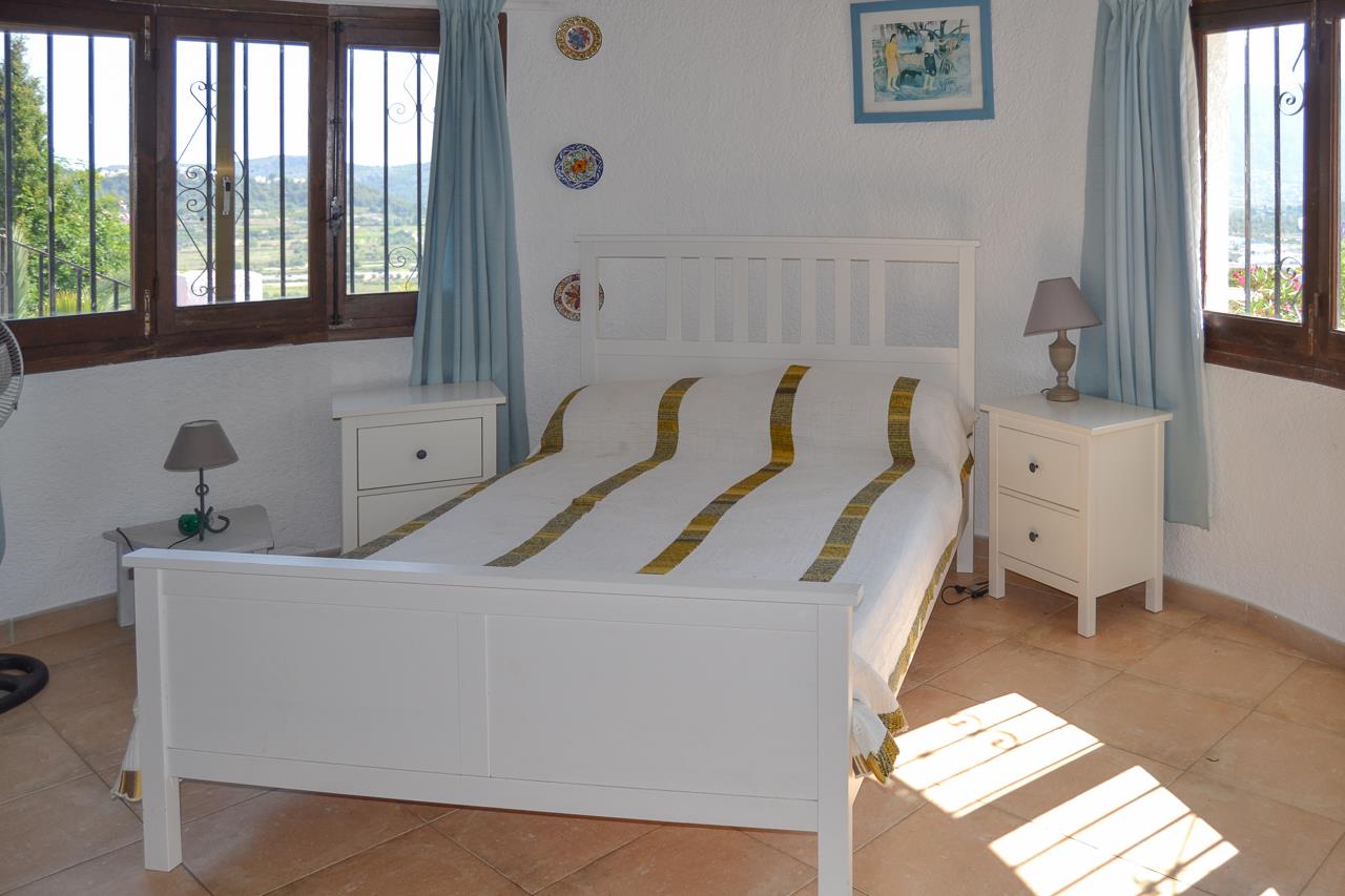 Bright bedroom featuring a white wooden bed frame, terracotta tile flooring, and windows with dark wood frames providing natural light and valley views.