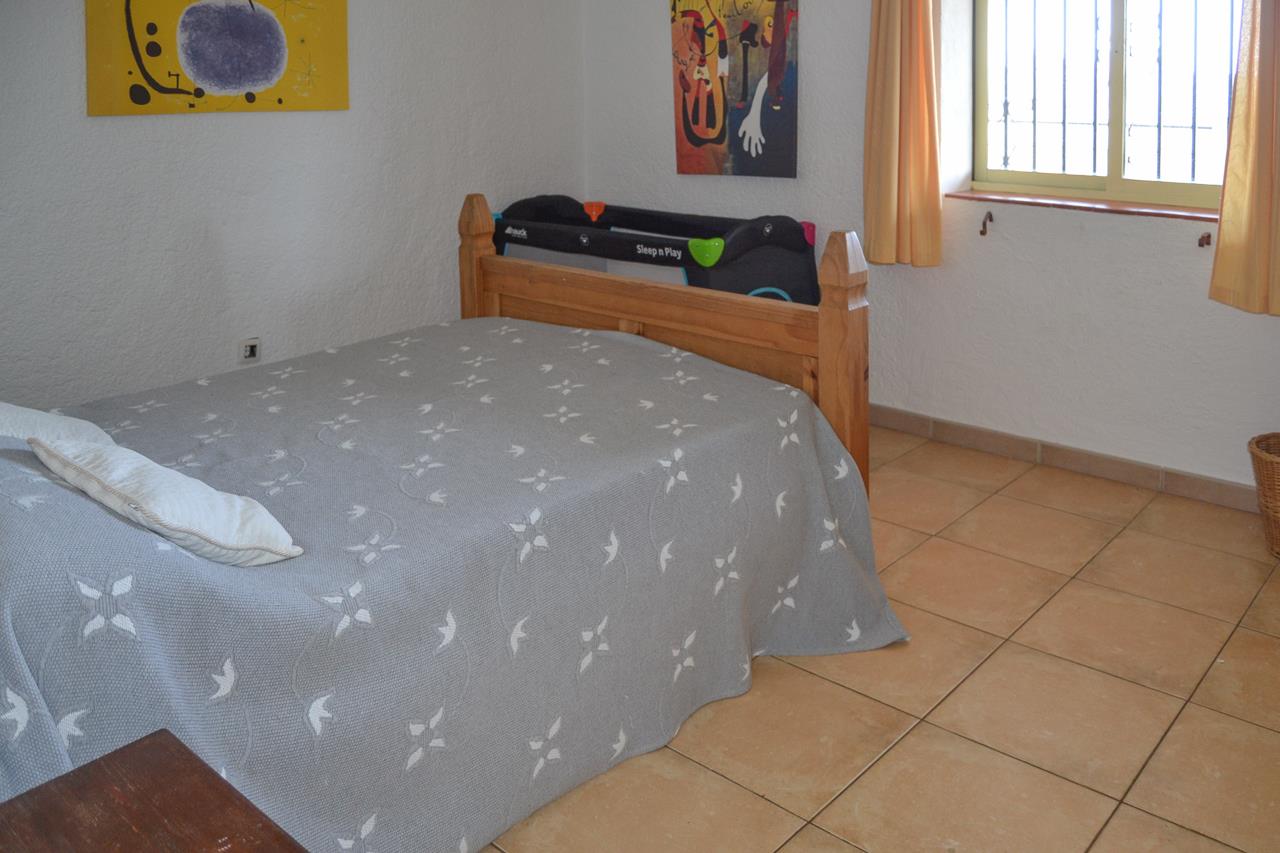 Bright bedroom featuring a wooden double bed, portable baby cot, and terracotta tile flooring. The room includes a window with security bars and colorful wall art.