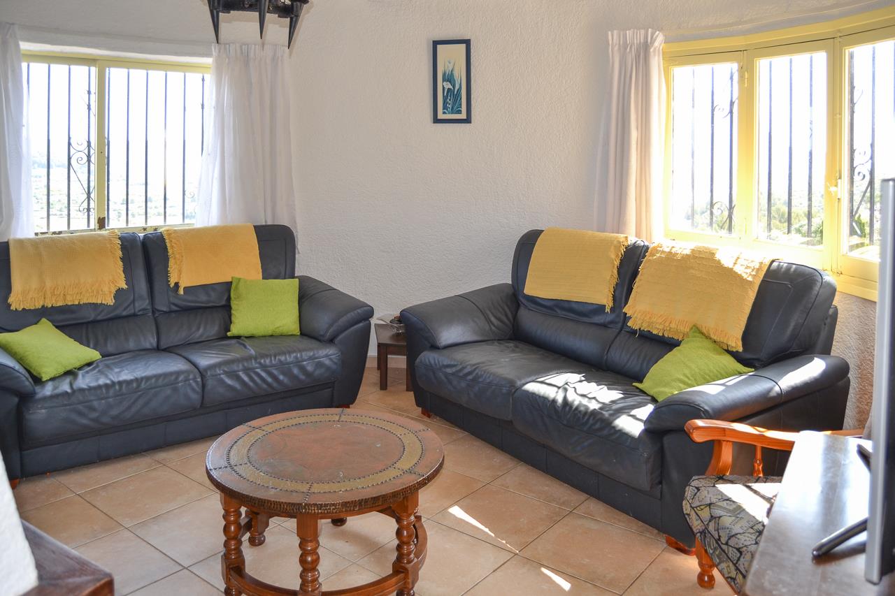 Bright living area featuring dark leather sofas, a carved wooden coffee table, and tiled flooring. Large windows with security bars provide ample natural light.