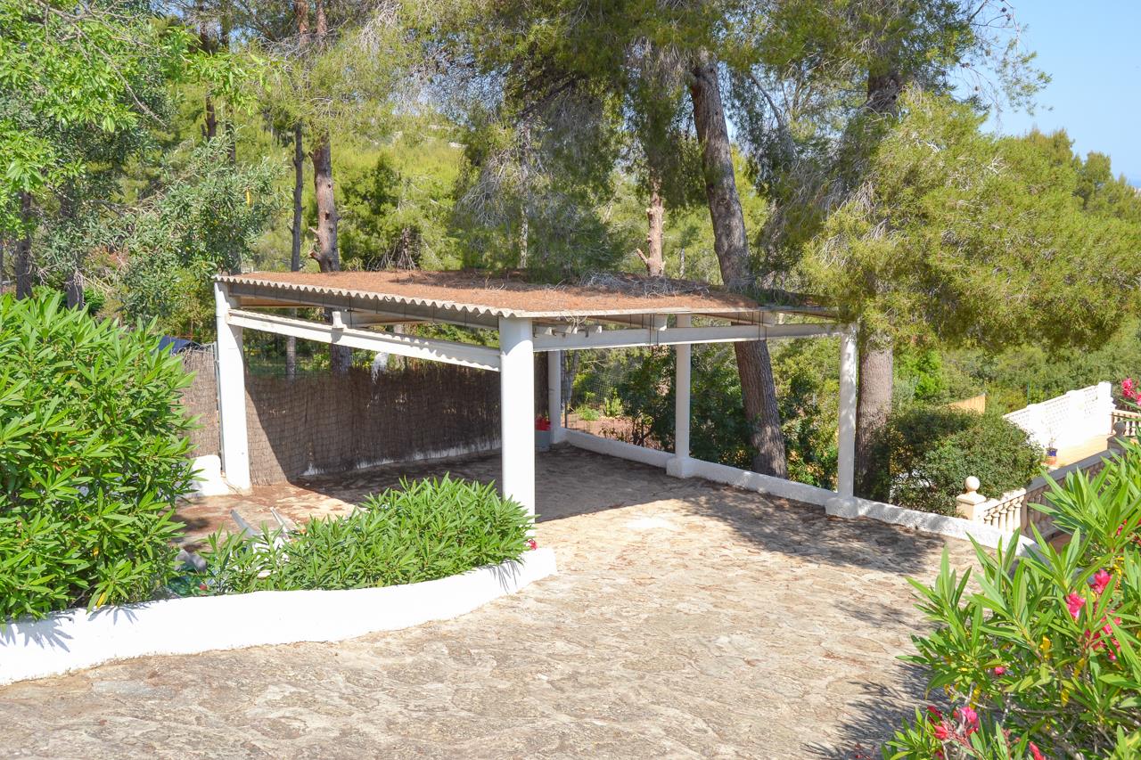 Covered parking area featuring a white structure and stone-paved driveway, nestled among mature pine trees and lush Mediterranean greenery.