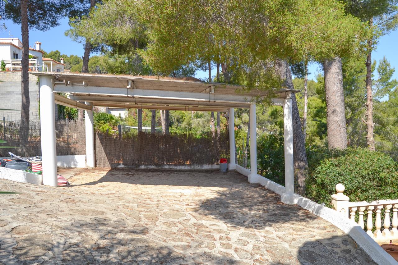 Covered parking space with white support pillars and natural stone paving, surrounded by mature pine trees on a sloped plot.