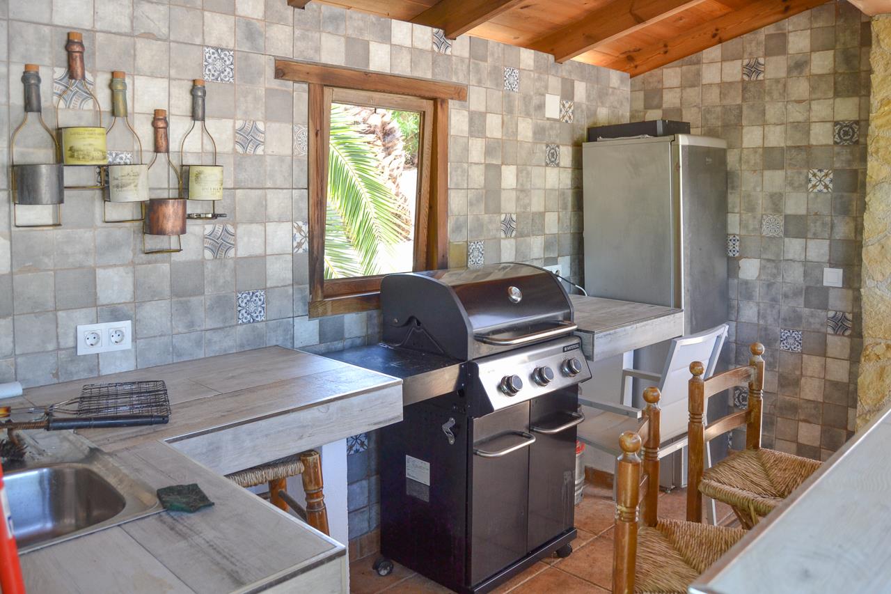 Covered summer kitchen featuring a gas grill, tiled countertops, and rustic wall mosaics. The space includes wooden ceiling beams, a sink, and views of Mediterranean greenery.
