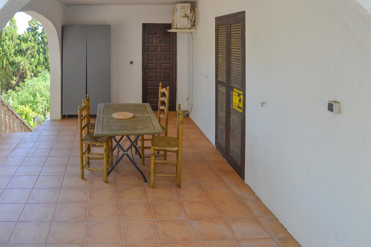 Covered terrace featuring terracotta floor tiles, a wooden dining set, and traditional dark wood doors set within a white arched structure.