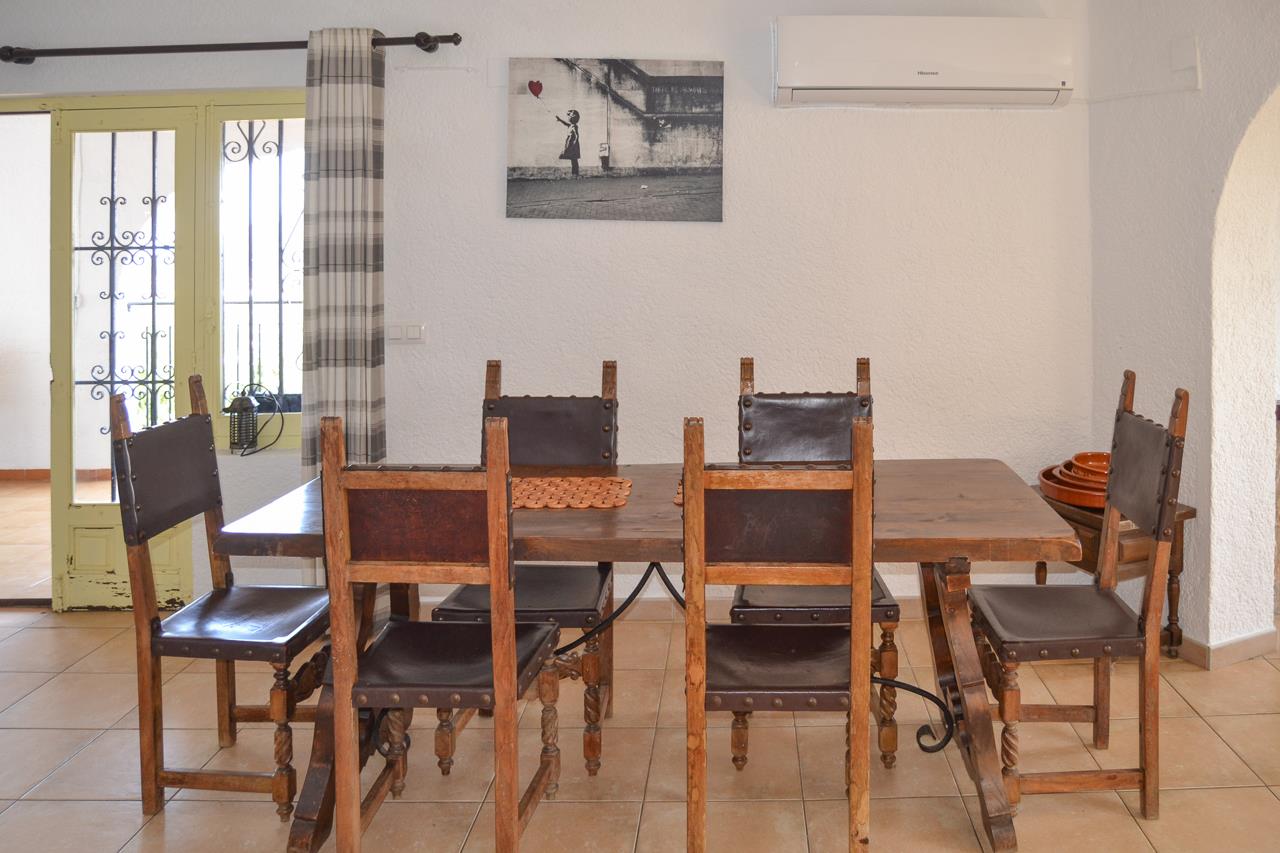 Dining area with a solid wood table and rustic leather chairs. Features include a wall-mounted air conditioning unit and traditional ceramic tile flooring.