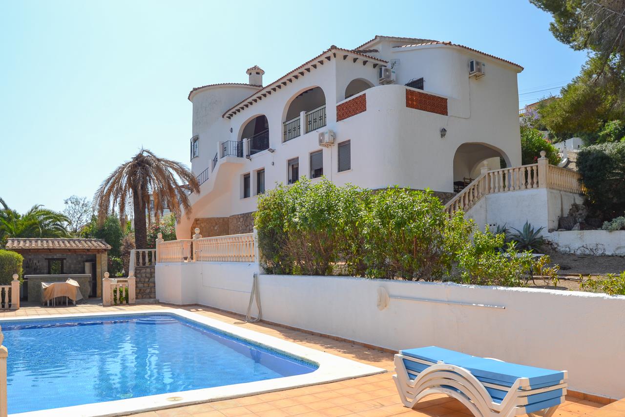 Exterior view of the villa featuring a large swimming pool, sun terraces, and Mediterranean architecture with arched verandas and stone accents.