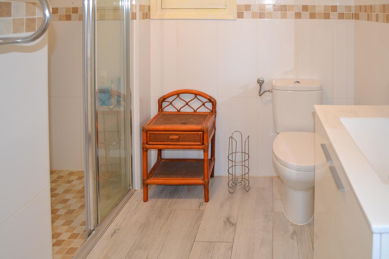Functional bathroom with a glass shower enclosure, white toilet, and rattan side table. Features light wall tiles with a mosaic border and wood-effect ceramic flooring.