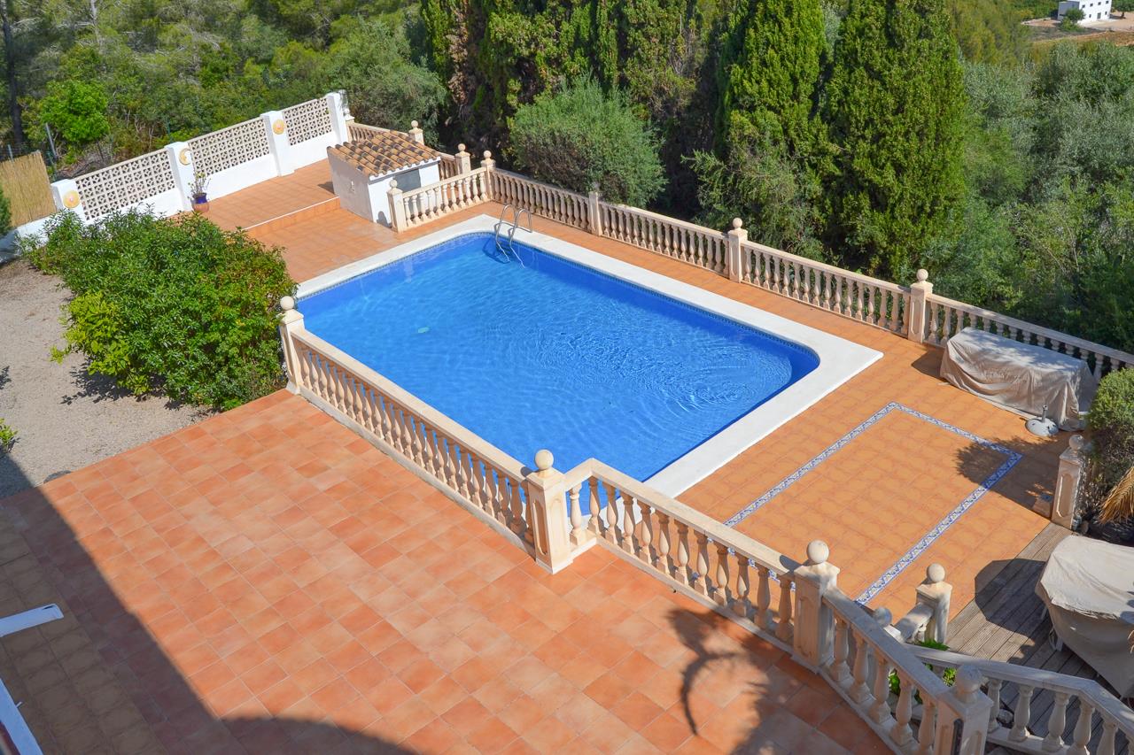 Large rectangular swimming pool featuring white stone coping, a terracotta tile terrace, and a classic stone balustrade, bordered by mature Mediterranean trees.