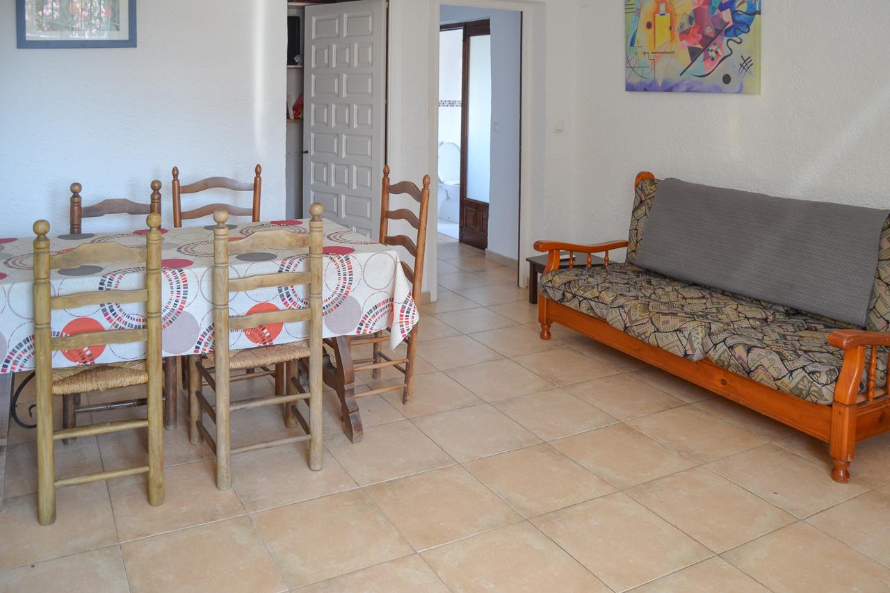 Living and dining area featuring ceramic tile flooring, a rustic wooden table set, and a wooden sofa. The space leads to a hallway and bathroom.