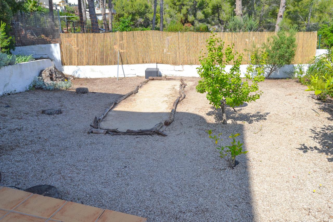 Low-maintenance gravel garden featuring a rustic petanque court bordered by natural logs, enclosed by a bamboo privacy screen and Mediterranean trees.