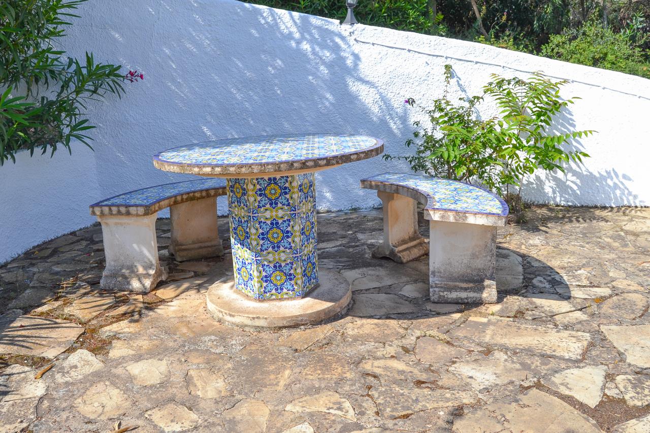 Outdoor seating area featuring a round table and curved benches decorated with traditional blue and yellow tiles, set on a natural stone paved terrace.