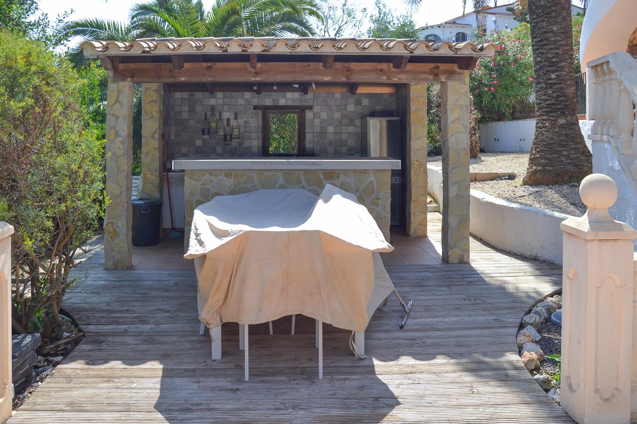 Outdoor summer kitchen featuring a stone-clad bar, masonry pillars, and a tiled roof over a wooden deck, set within a lush Mediterranean garden.