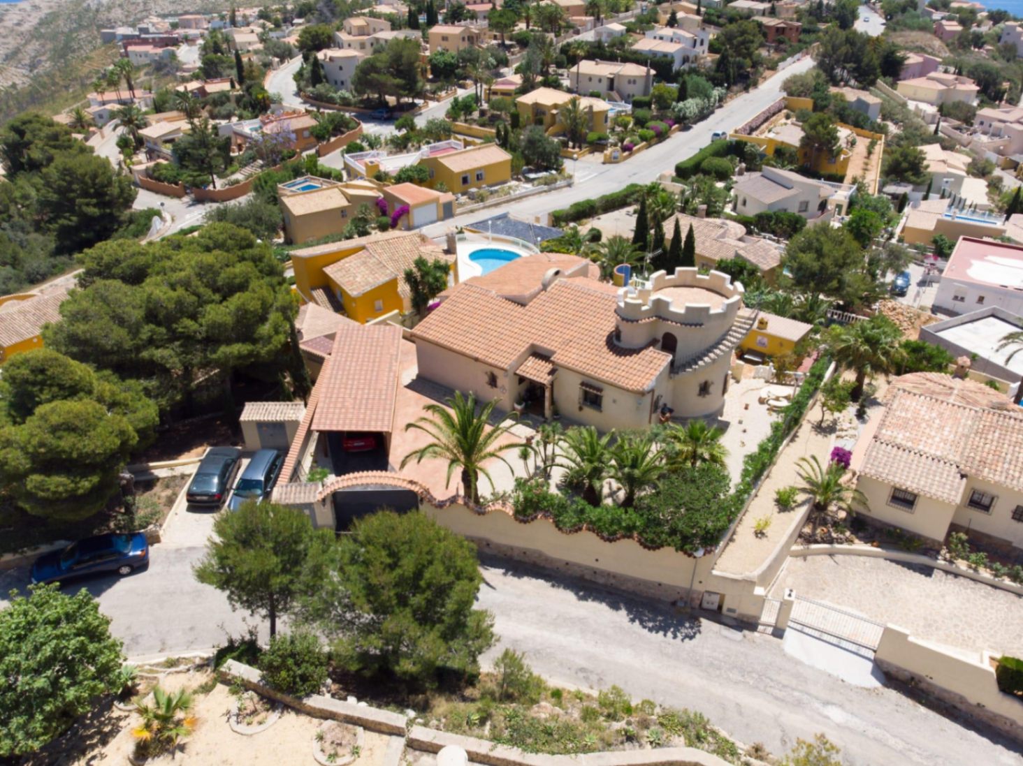 Aerial view of the villa featuring a distinctive crenellated tower, terracotta tiled roof, and Mediterranean garden with palm trees on an elevated plot.