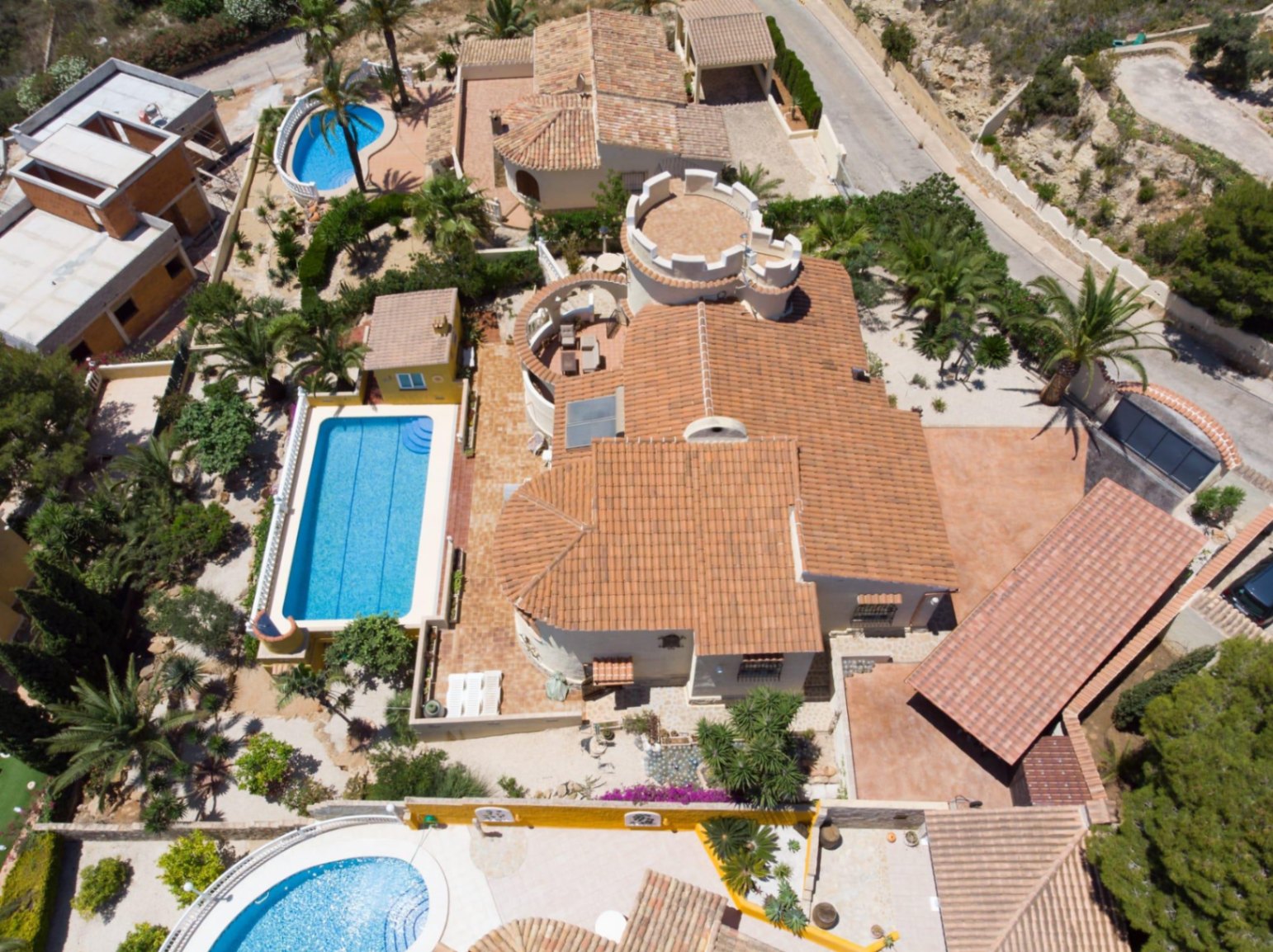 Aerial view of the villa featuring its terracotta tile roof, distinctive crenellated circular tower, and a rectangular pool set within Mediterranean gardens.