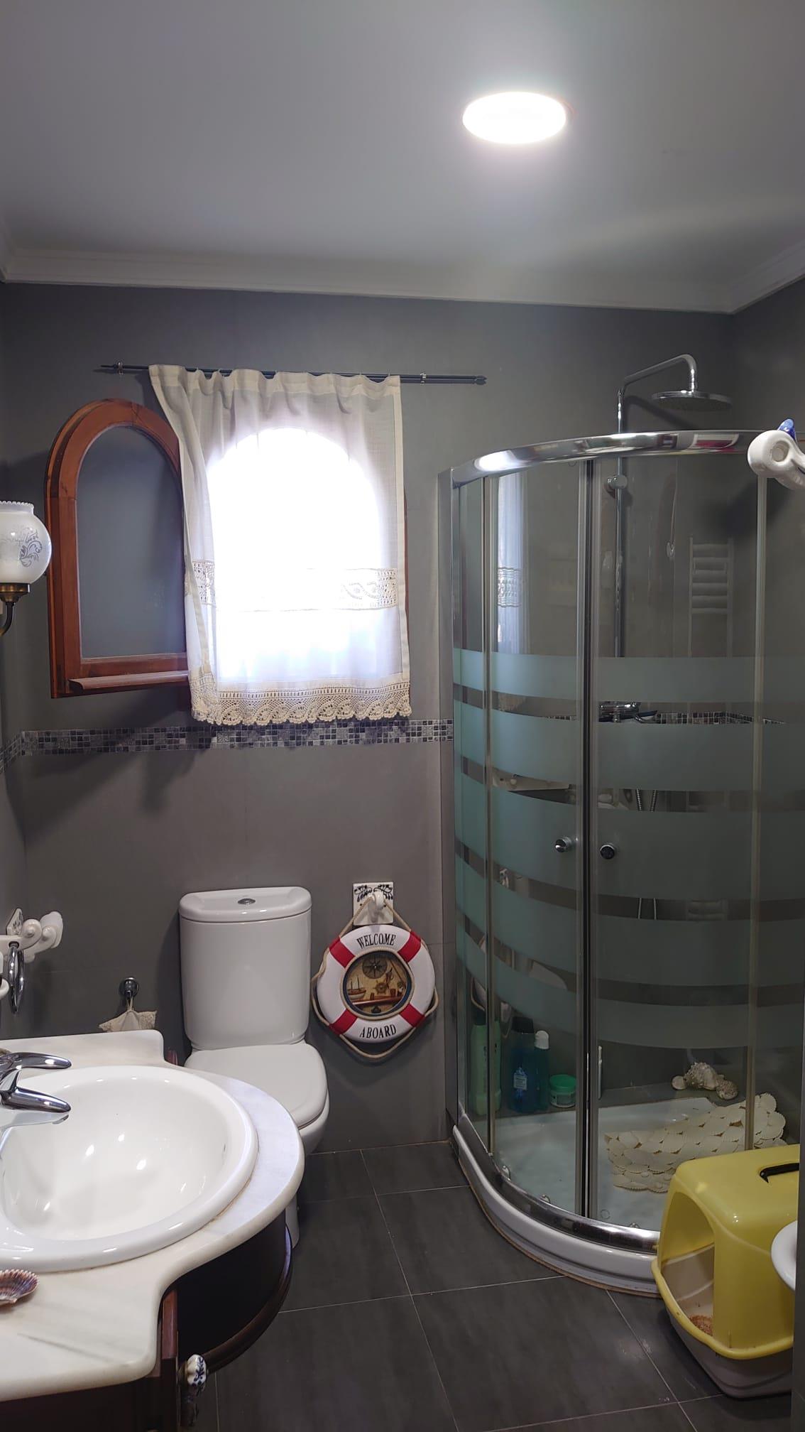 Bathroom featuring a curved glass corner shower, grey tiled walls, and a marble vanity. It includes an arched wooden window frame and nautical-themed decor.