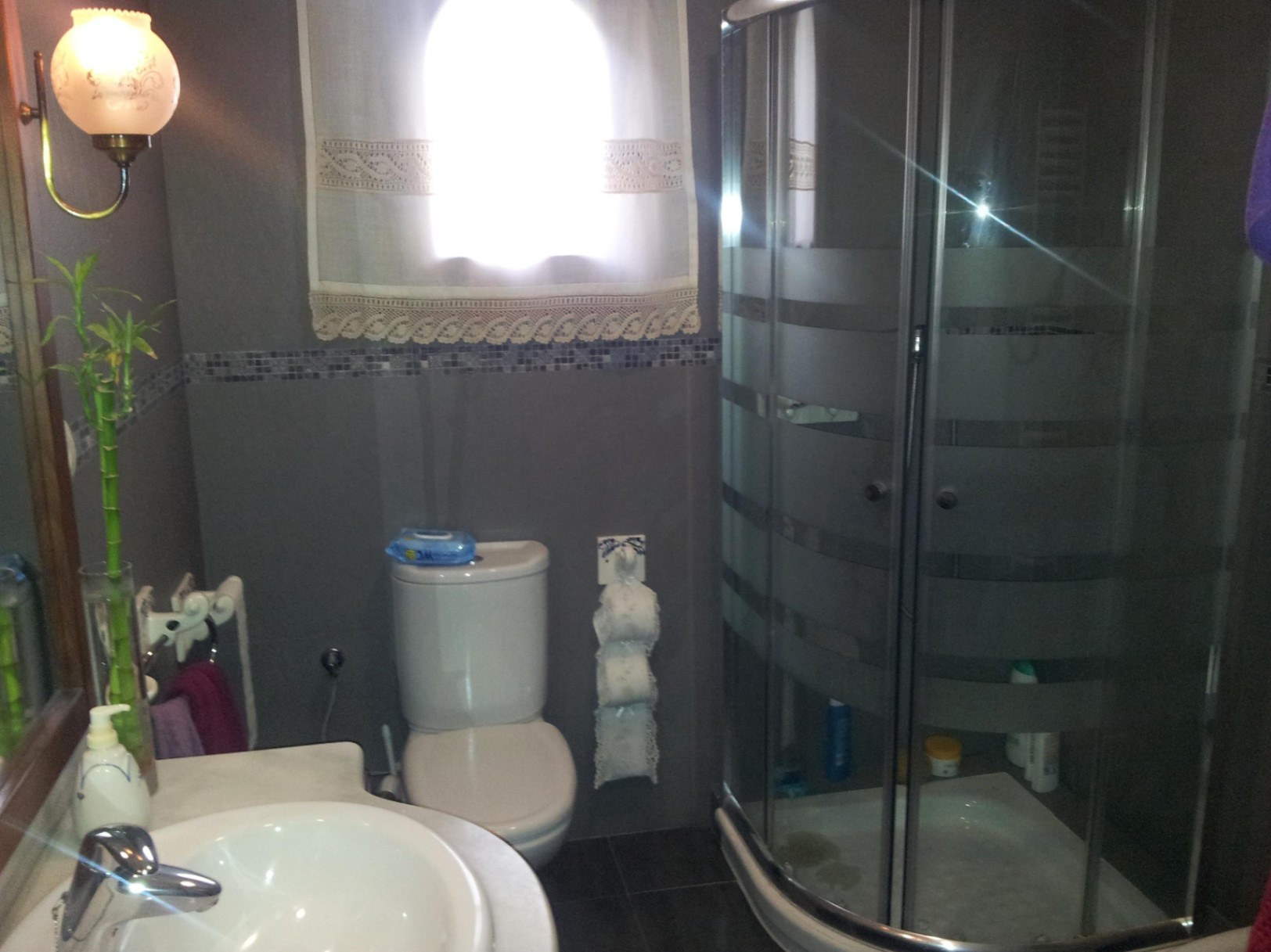 Bathroom featuring a curved glass corner shower, grey tiled walls with a mosaic border, and white ceramic fixtures illuminated by natural light.