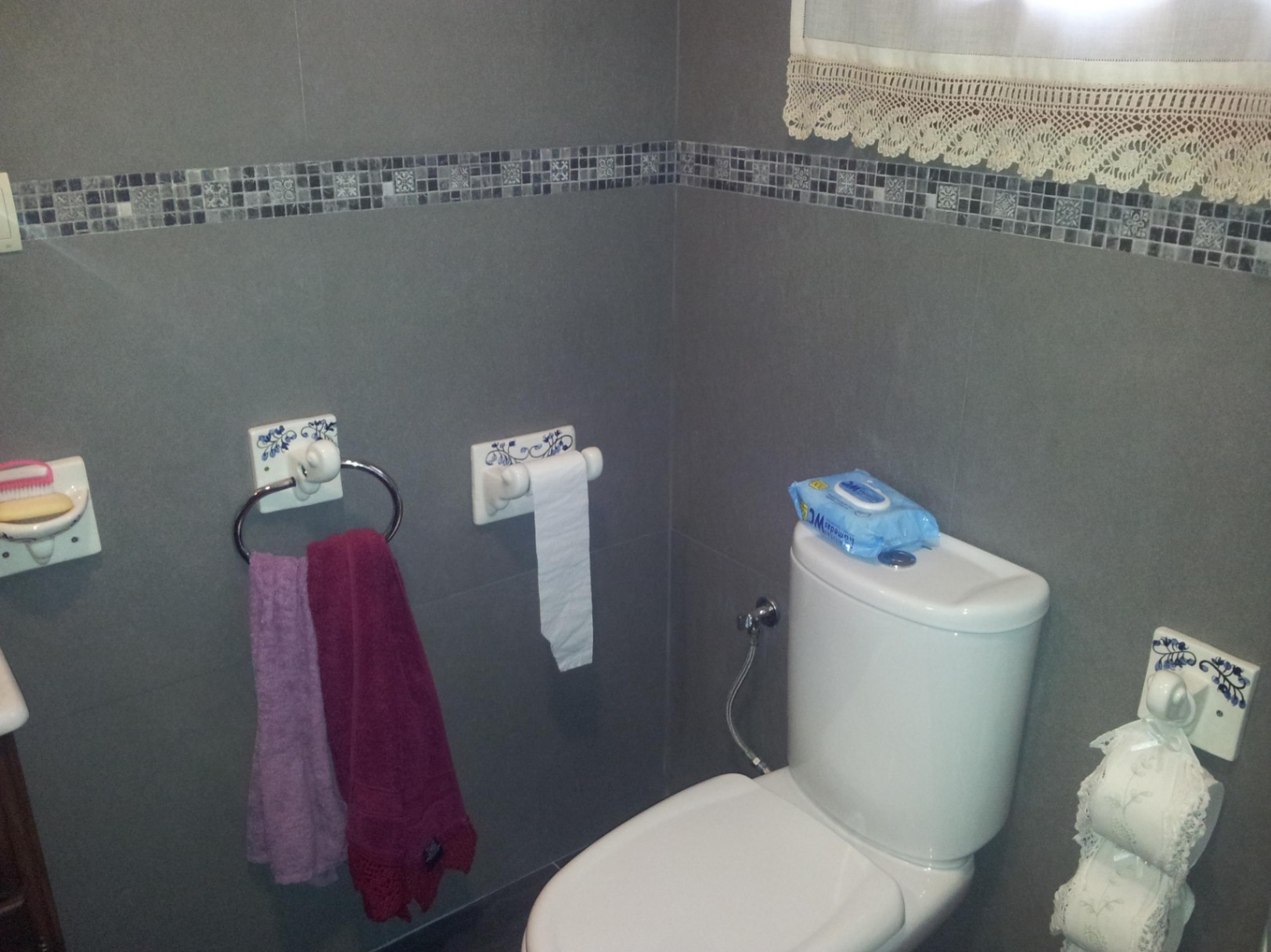 Bathroom featuring grey wall tiles with a mosaic border, white ceramic toilet, and traditional ceramic accessories with blue floral patterns.