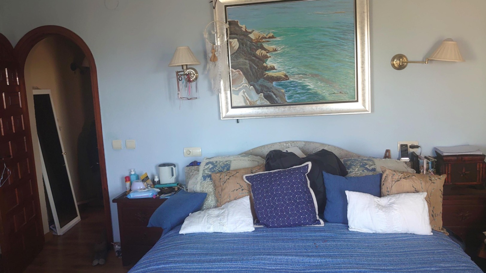 Bedroom featuring a bed with blue linens, a large coastal painting in a silver frame, and twin wall lamps. An arched doorway leads to the adjacent room.